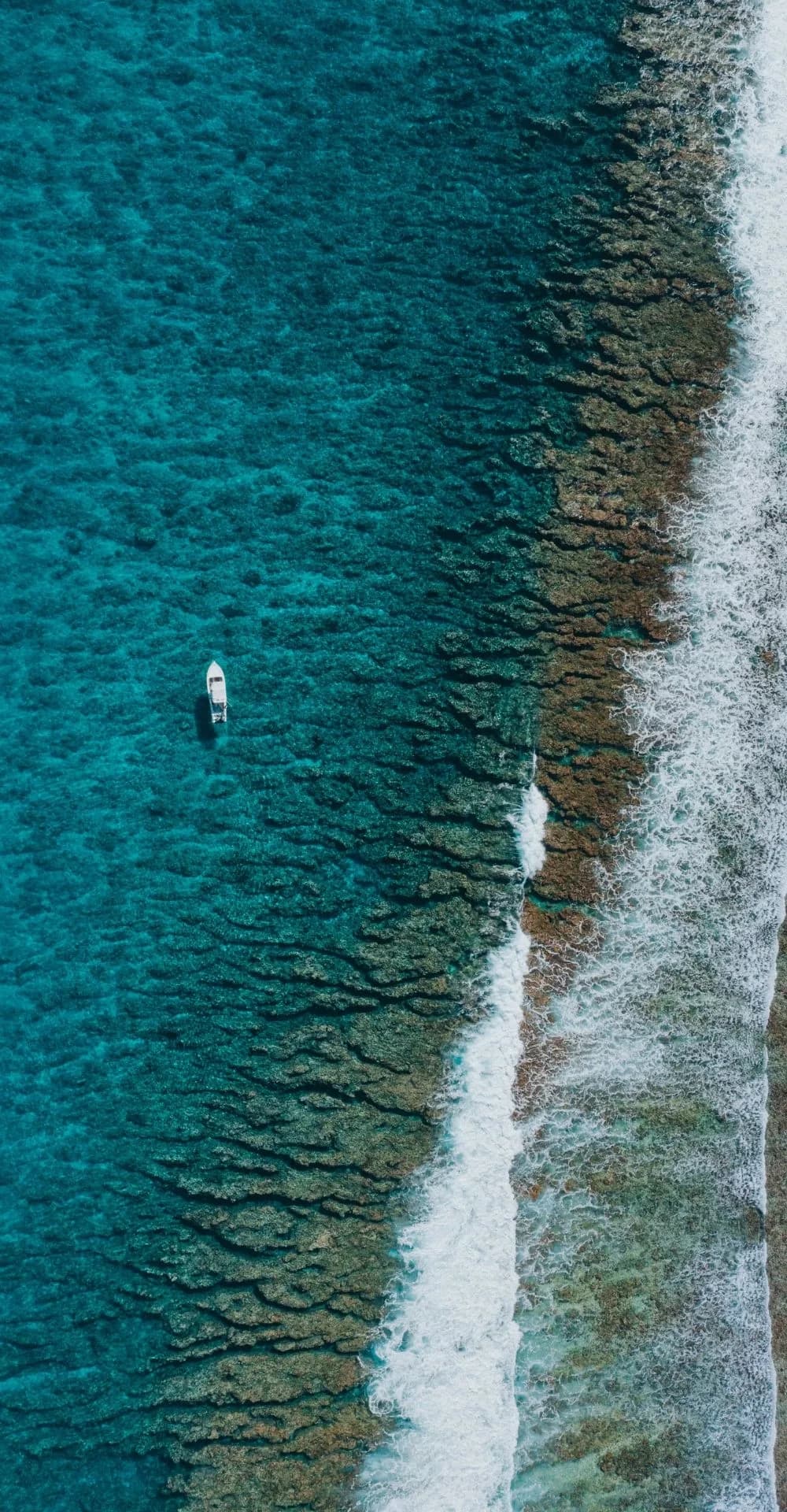 A small white boat on turquoise water beside a reef with breaking waves, seen from the air