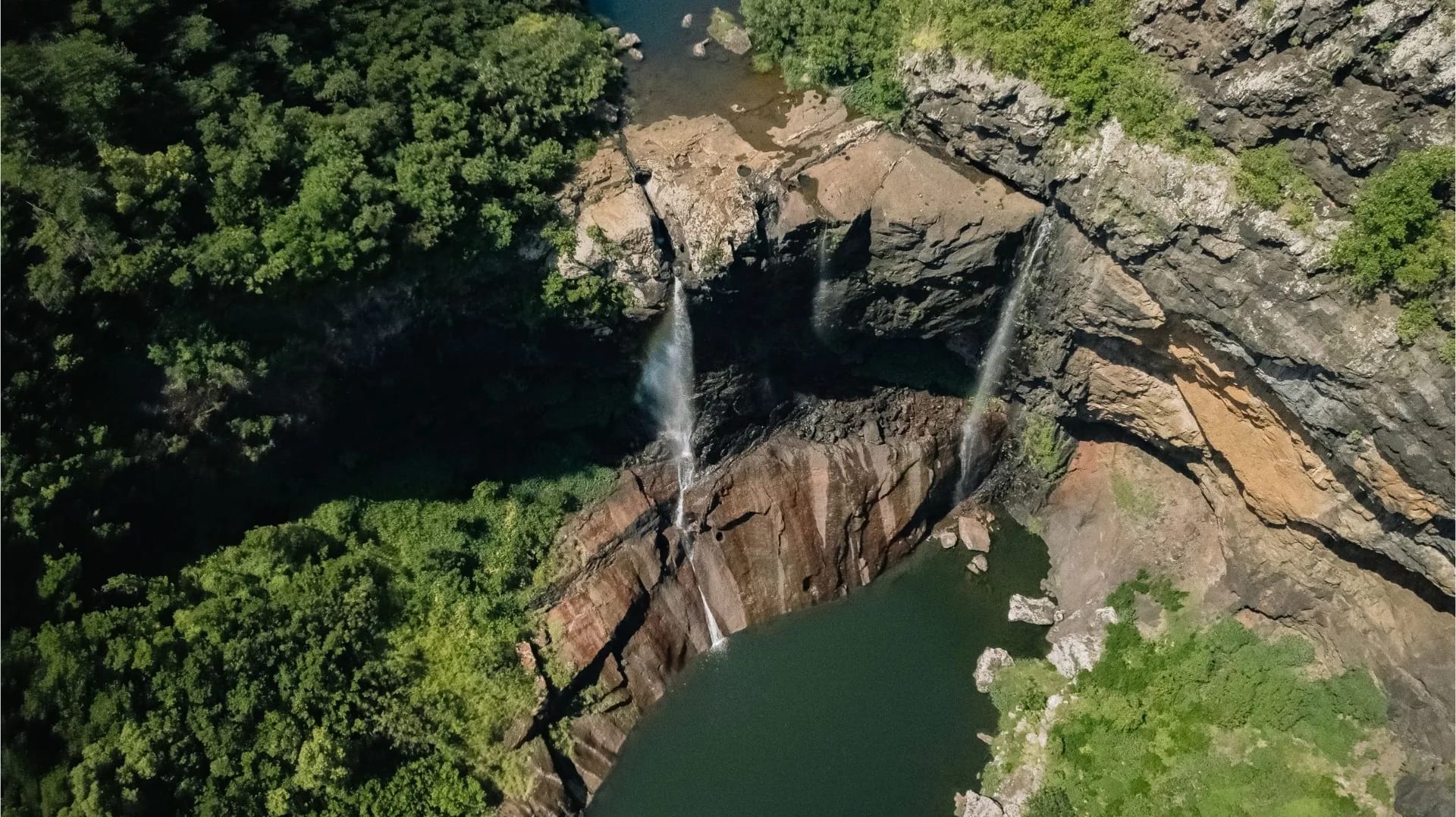 A waterfall and pool between rocky cliffs and green vegetation at Tamarind Falls, seen from above. Photograph from Yanature.
